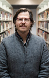 white man in glasses standing between bookshelves