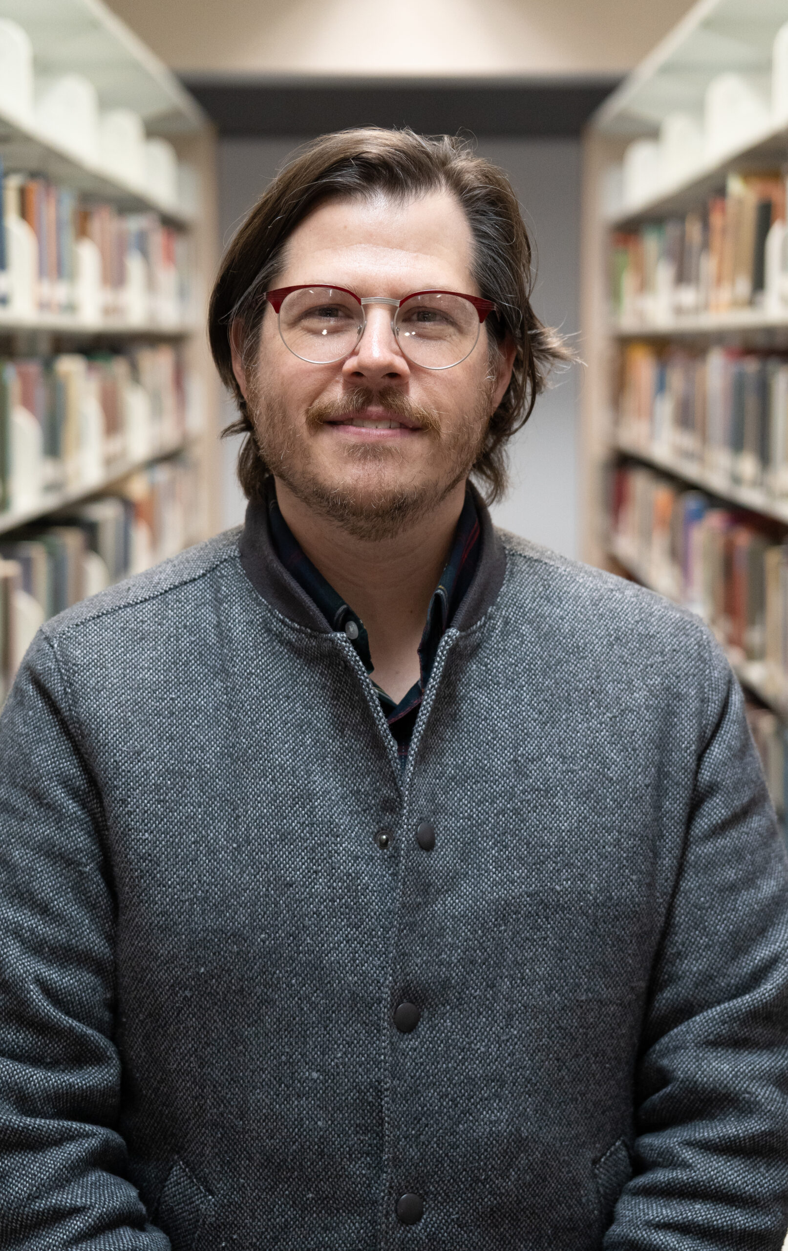 white man in glasses standing between bookshelves